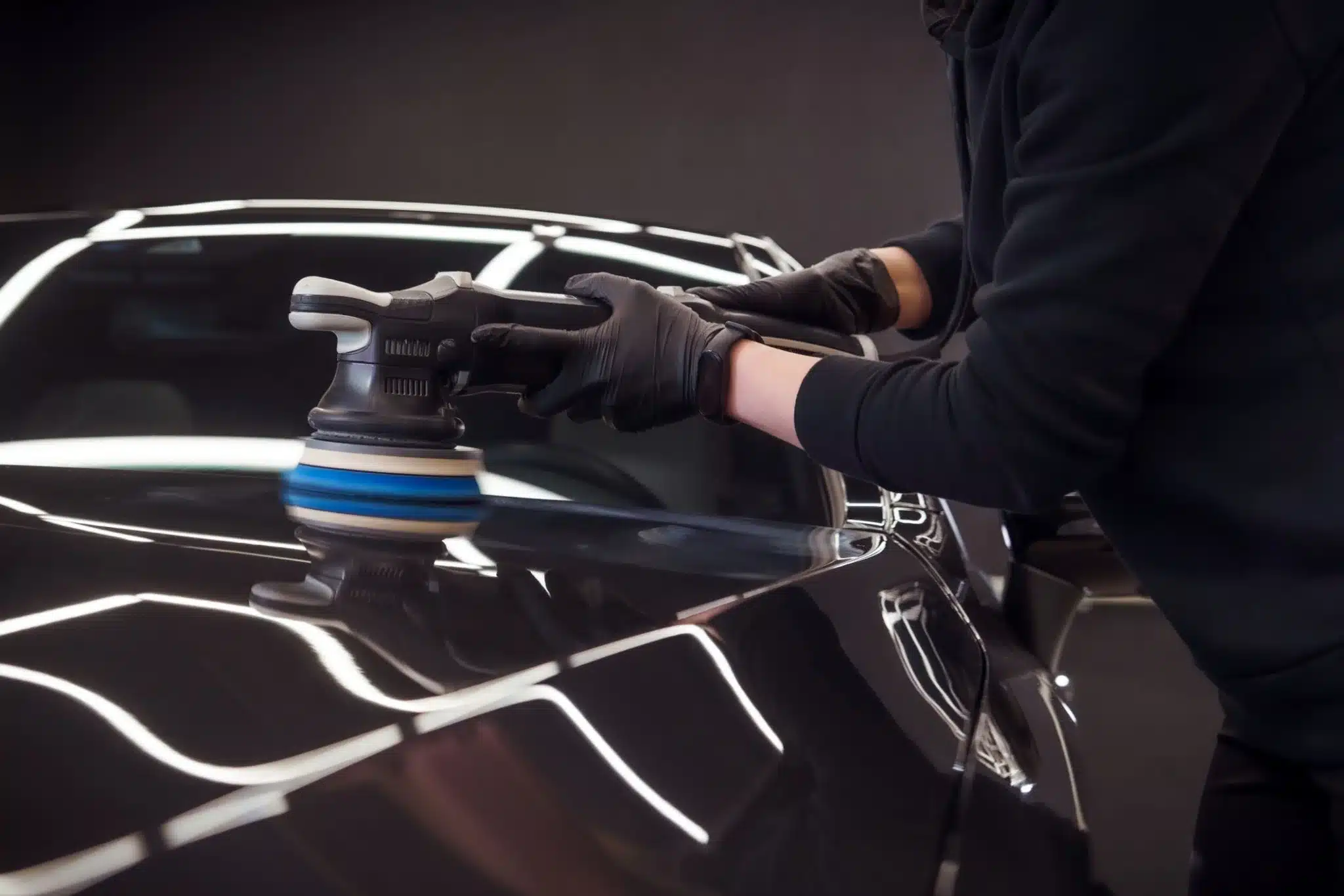 A professional detailer in gloves using a dual-action machine polisher on a dark car bonnet under bright workshop lighting